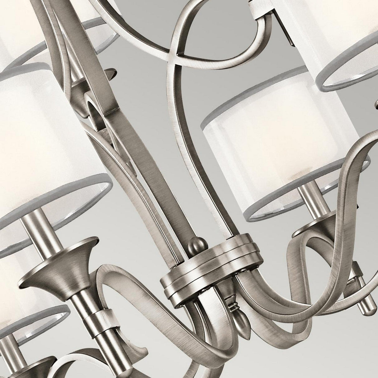 The Lacey 9 Light Chandelier - Silver is captured in a close-up view, highlighting its curved metallic arms with a silver finish and cylindrical white lampshades set against a neutral background. The chandelier's intricate metalwork displays smooth, flowing lines that embody an elegant dual-shade design.