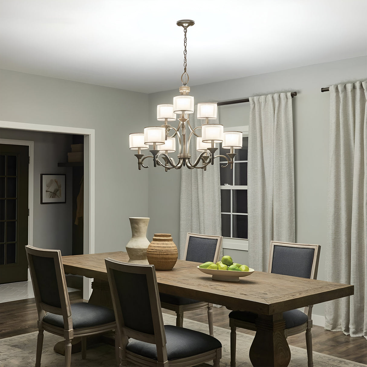 In a dining room featuring a wooden table with six chairs, the Lacey 9 Light Chandelier - Silver casts a warm glow overhead. Vases and a bowl of green apples elegantly adorn the table, while light gray walls and curtains frame the space, contributing to the serene ambiance of this chic design.