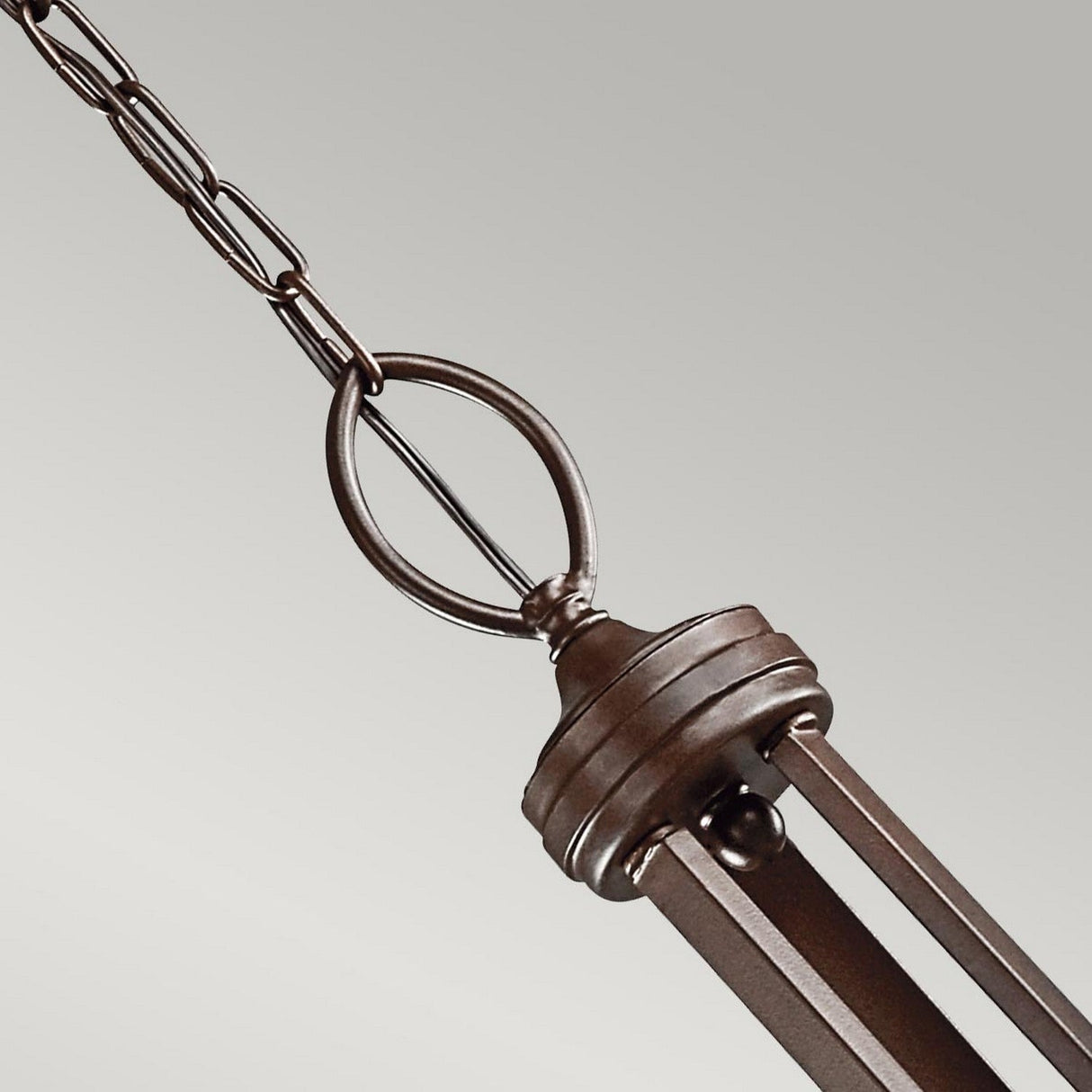 A close-up of the Lacey 9 Light Chandelier - Bronze showcases its metallic finish. The design, featuring a subtle bronze hue, includes interconnected metal rods and rings suspended from a chain, reminiscent of a classic chandelier against a plain background.