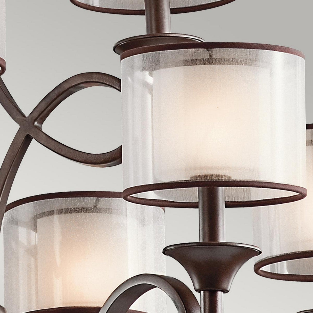 Take a close look at the Lacey 9 Light Chandelier in bronze, showcasing an exquisite double-shade design with frosted cylindrical glass shades. Its elegant metal frame is adorned with curved details, and each light provides a soft, warm glow against a neutral background.