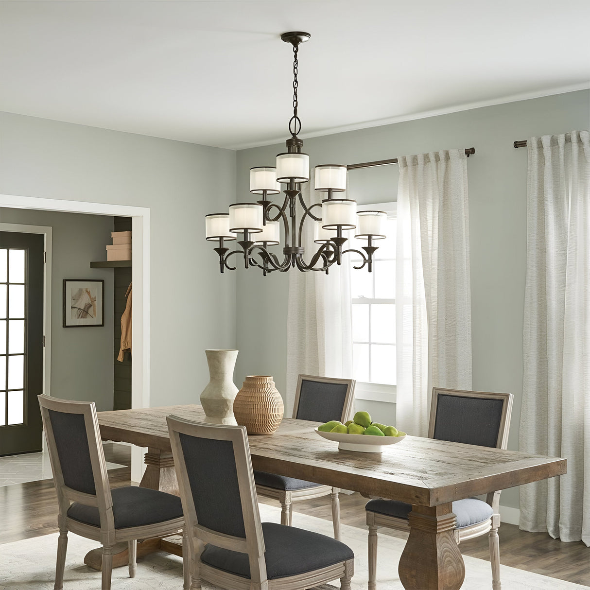 A dining room featuring a rustic wooden table and four cushioned chairs is beautifully accented by a cluster of decorative vases. Hanging above, the Lacey 9 Light Chandelier with a bronze finish adds elegance to the space. Sunlight pours through sheer curtains, creating a warm and inviting atmosphere.