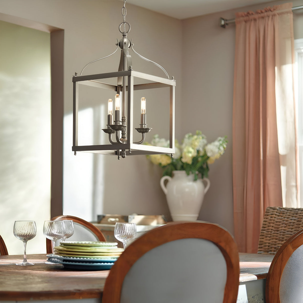 The dining room showcases the Larkin 3 Light Medium Chandelier in brushed nickel, with its open cage design illuminating a warm glow over the wooden table set elegantly with glassware and stacked plates. In the background, sheer curtains frame a large vase filled with white flowers.
