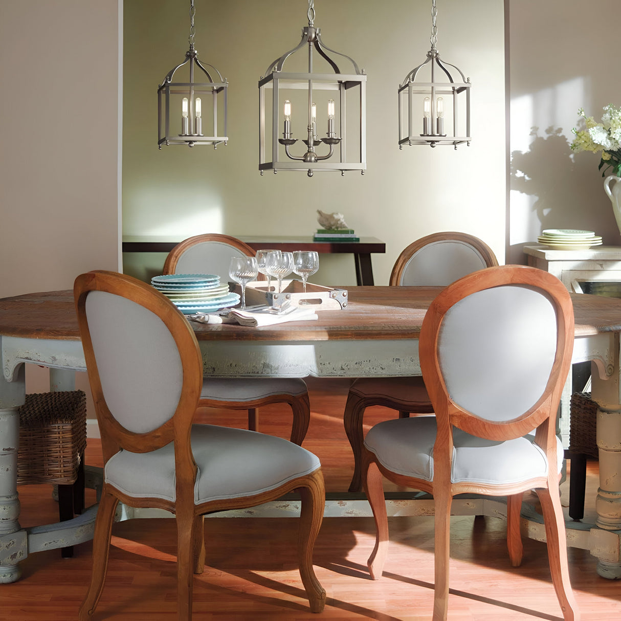 A cozy dining area showcasing an oval wooden table surrounded by light blue cushioned chairs, with the glow of a Larkin 3 Light Medium Chandelier in brushed nickel illuminating the space. In the background, a shadowed room containing a console table and decorative items adds depth to this inviting scene.