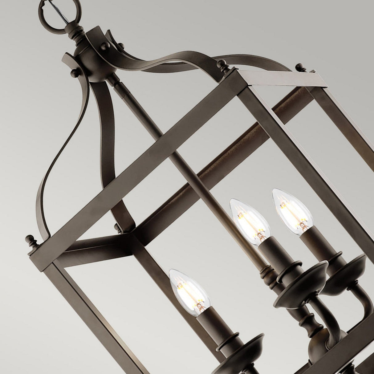 A detailed view of the Larkin 3 Light Medium Chandelier - Olde Bronze reveals its metal structure, featuring an open cage design and a rectangular frame that holds four candle-shaped bulbs. With its sophisticated Olde Bronze finish set against a neutral backdrop, this chandelier masterfully combines rustic and elegant elements.