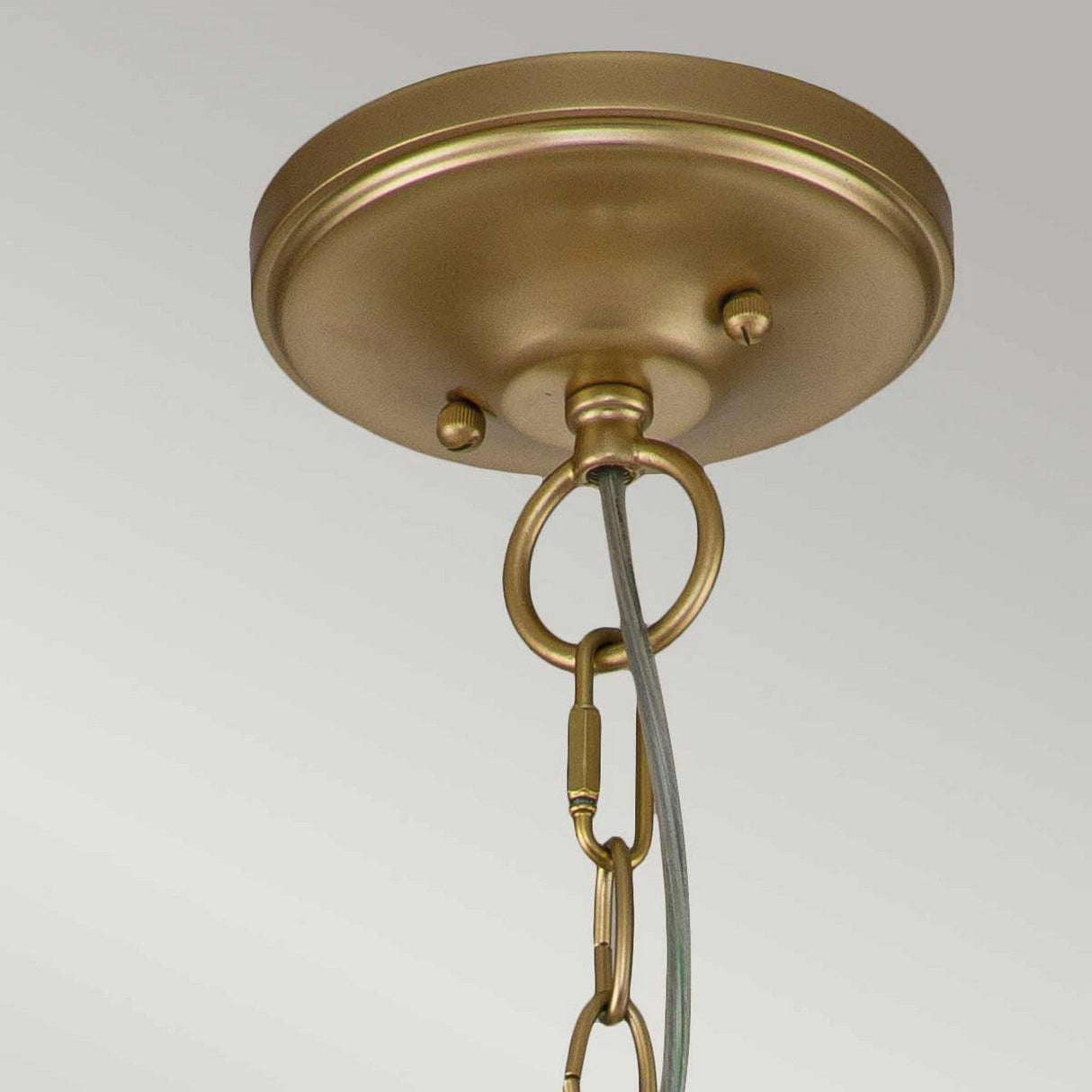 A close-up of the Larkin 3 Light Medium Chandelier in natural brass shows its circular ceiling mount. The metal hook is attached to a linked chain, perfect for hanging a fixture. Its backdrop is plain and light-colored, reflecting the elegant simplicity of its open cage design.