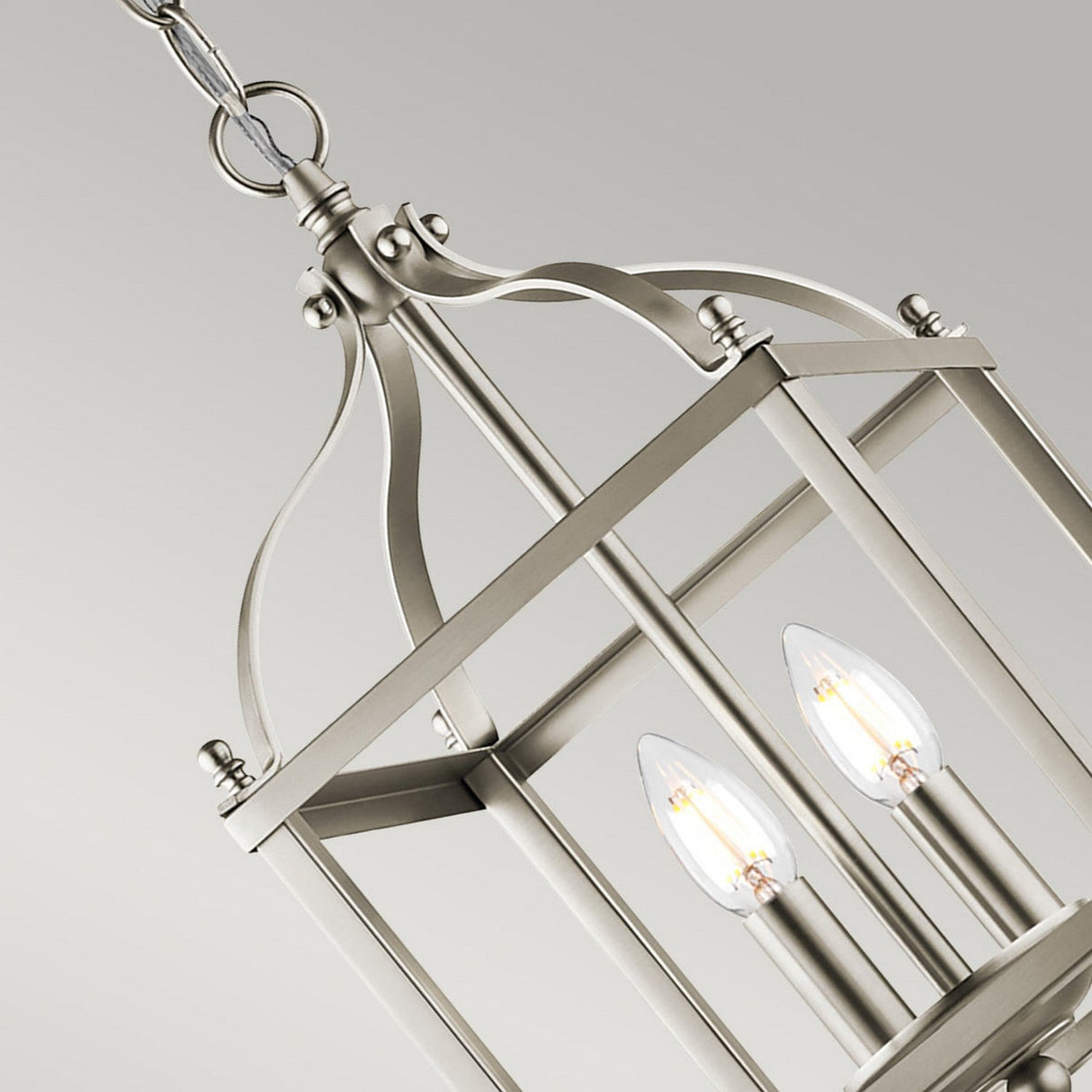 A close-up of the Larkin 2 Light Small Chandelier in brushed nickel, highlighting its modern open cage design. The geometric frame elegantly displays two exposed bulb lights, suspended from a chain against a neutral, light background.