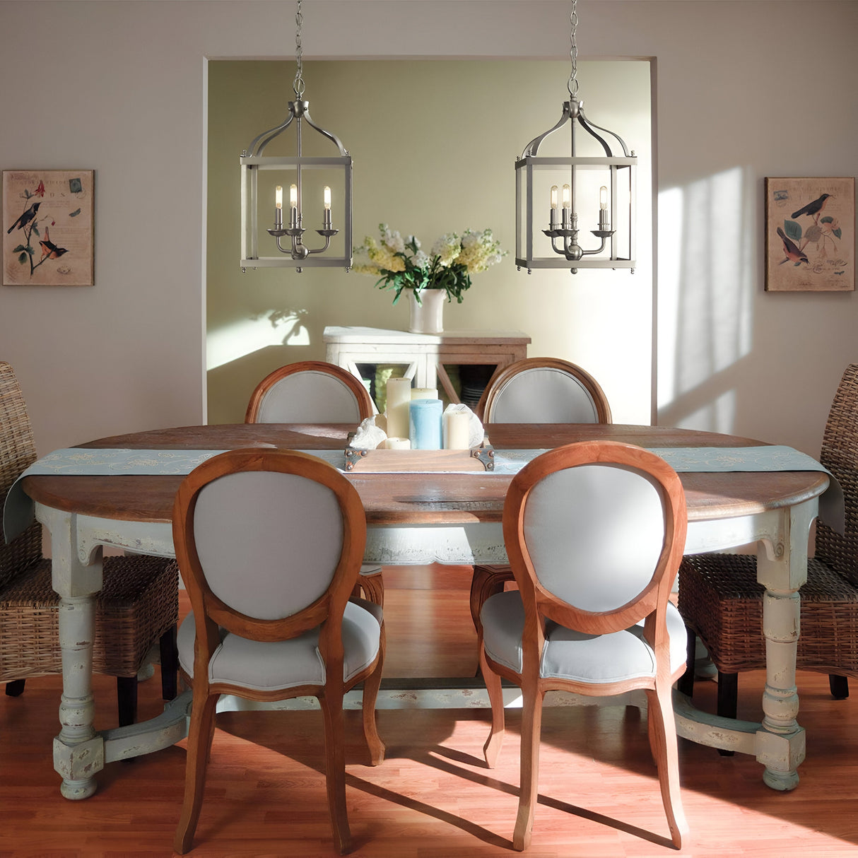 A cozy dining room features an oval wooden table, elegantly arranged with candles and a floral centerpiece, surrounded by four upholstered chairs. Overhead, two Larkin 2 Light Small Chandeliers in Brushed Nickel provide a warm glow, while bird artwork decorates the walls. The space is filled with natural light.