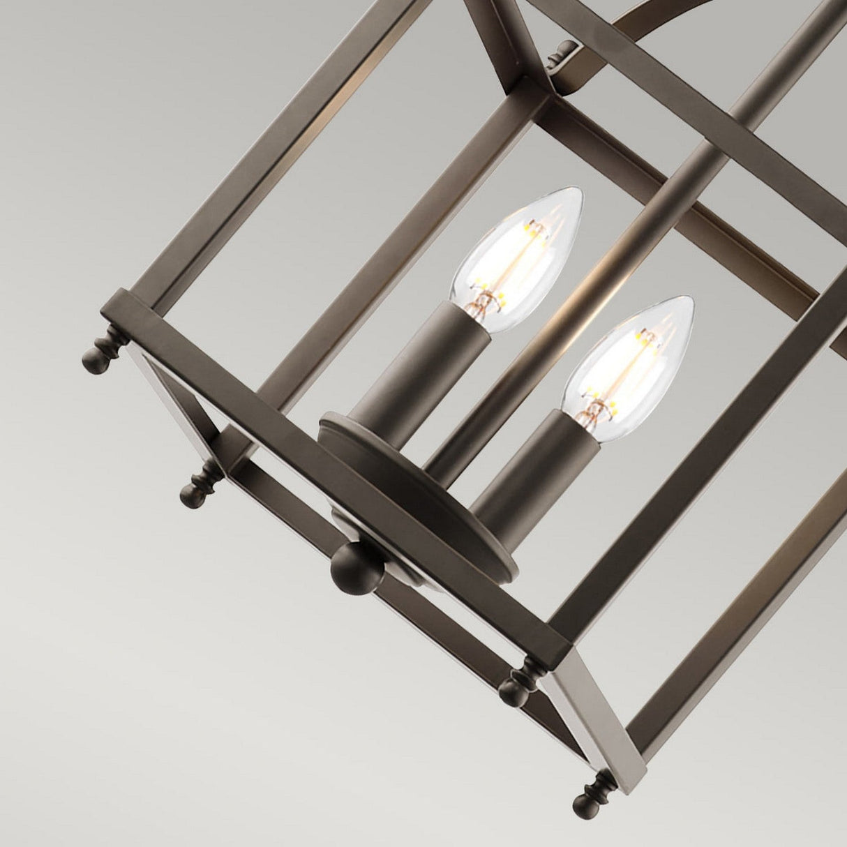 A detailed view of the Larkin 2 Light Small Chandelier showcases its modern, rectangular metal frame in an open cage design, featuring two exposed bulb-shaped lights. It is elegantly suspended in a minimalist style and finished in Olde Bronze against a plain gray background.