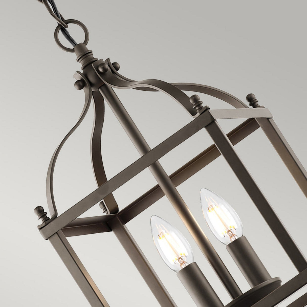 A close-up of the Larkin 2 Light Small Chandelier in a rich Olde Bronze finish features two exposed light bulbs within an intricate open cage design. Hanging elegantly from a chain against a neutral backdrop, this fixture beautifully combines modern and classic styles.