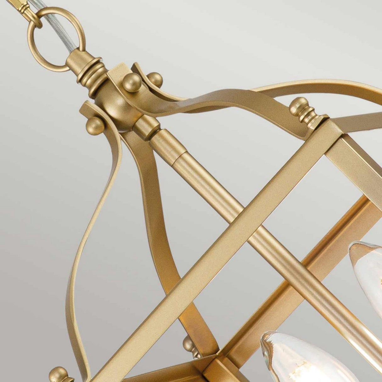 A close-up view of the Larkin 2 Light Small Chandelier in natural brass showcases its modern open cage design with geometric features and candle-style bulbs. The combination of curved and straight lines is accentuated by a circular attachment, set against a plain light gray backdrop.