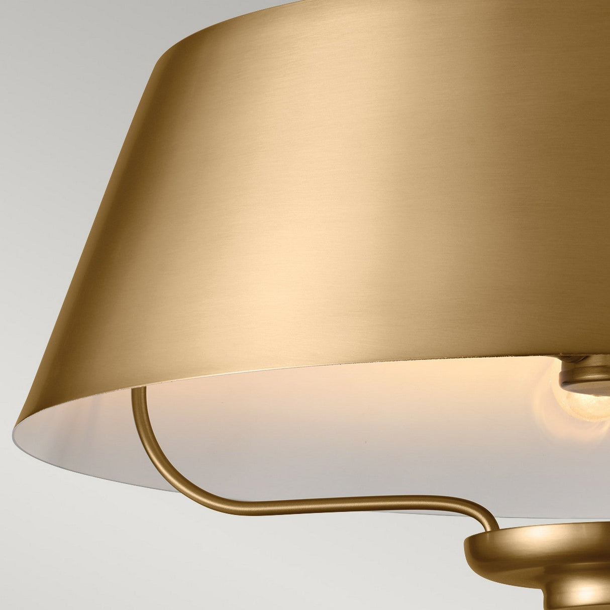 Close-up of the Luella 3 Light Duo-Mount Chandelier/Semi-Flush in brushed natural brass, featuring a modern, gold-colored finish with a sleek, metallic shade. The light bulbs inside emit a soft glow, illuminating the top part of the lamp's interior. This elegant lighting solution stands out against the neutral, light gray background.