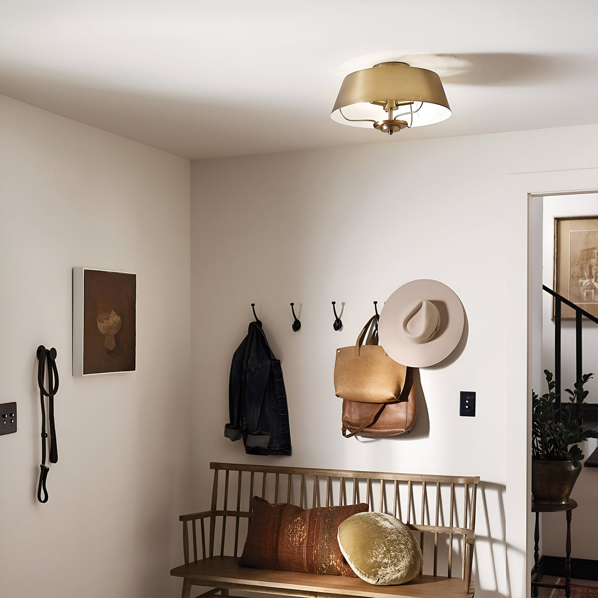 A cozy entryway showcases a wooden bench with pillows and a wall adorned with hooks holding jackets, bags, and a hat. Above, the Luella 3 Light Duo-Mount Chandelier/Semi-Flush in Brushed Natural Brass illuminates the space. A piece of artwork and a railing leading downstairs complete the scene.