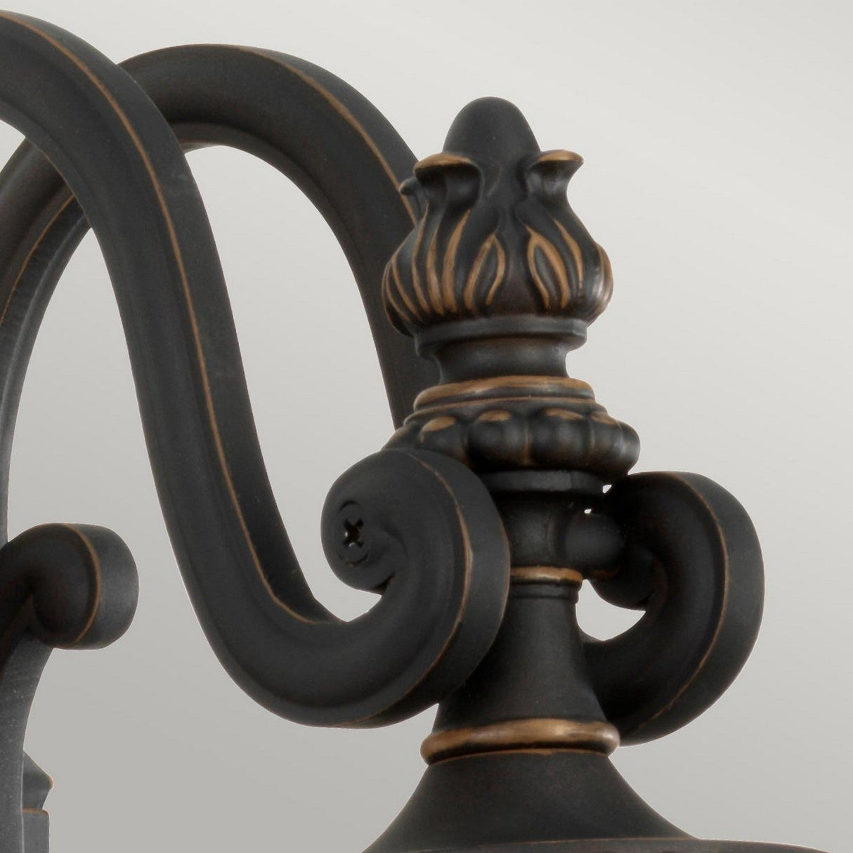 Close-up of the Luverne Outdoor 1 Light Medium Wall Lantern, highlighting its decorative metal scrollwork with a flame-shaped finial. The rubbed bronze finish is enhanced with subtle gold accents, displaying intricate detailing and a curved design against a light gray background, reminiscent of an elegant outdoor wall lantern.