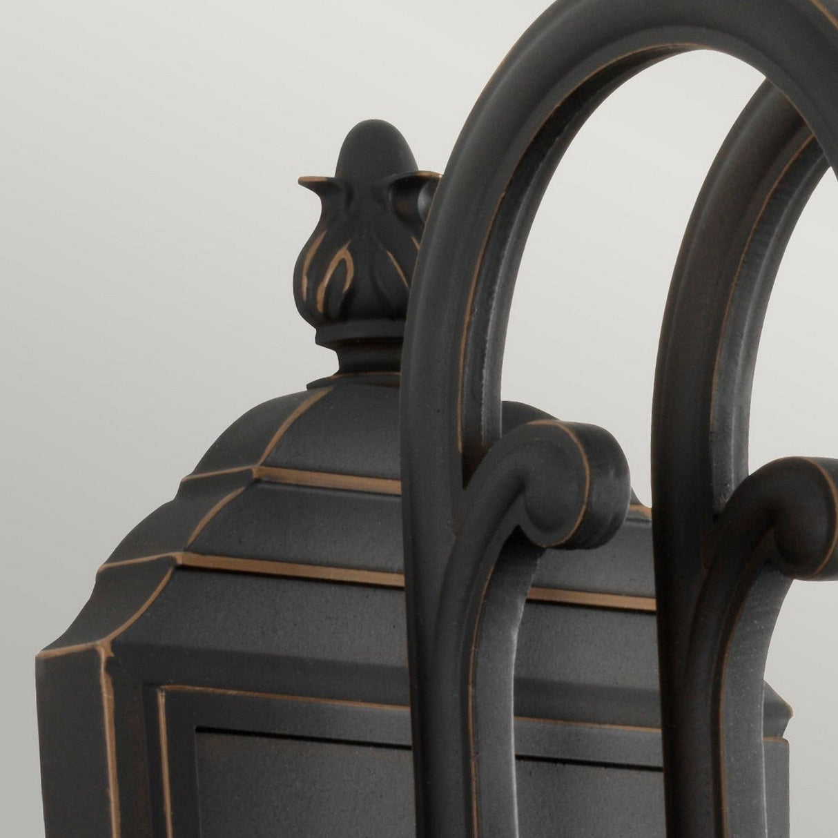 Close-up of the Luverne Outdoor 1 Light Medium Wall Lantern in rubbed bronze, showcasing its ornate scrolling metalwork and decorative finial atop a ribbed glass shade, all set against a light gray background.