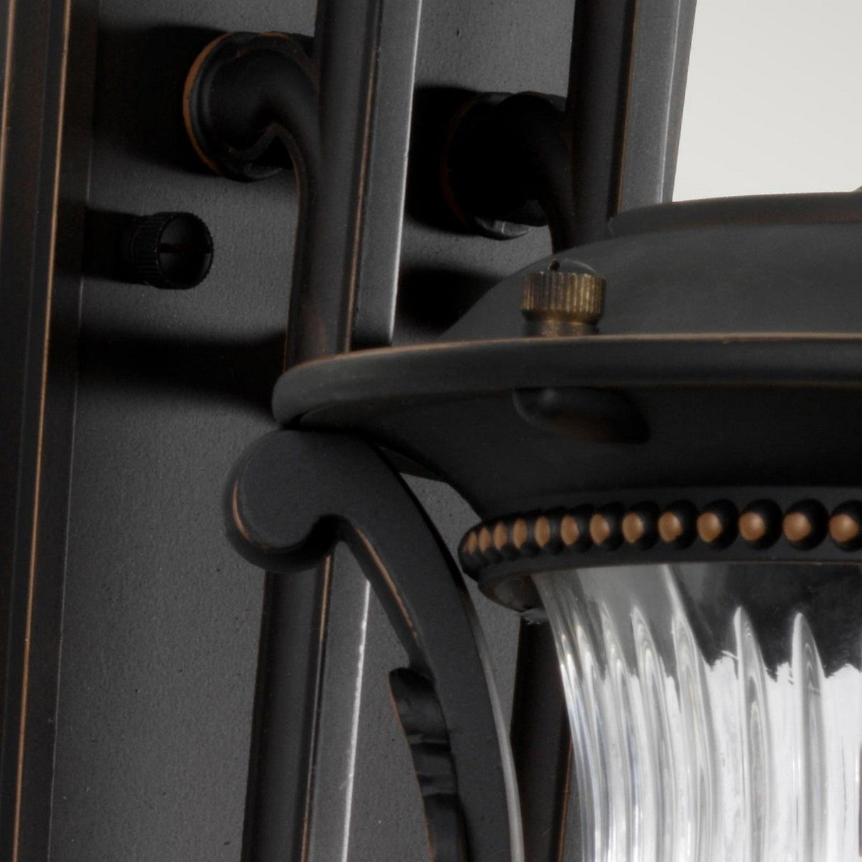 A close-up of the Luverne Outdoor 1 Light Medium Wall Lantern in rubbed bronze shows the detailed brackets and a clear, ribbed glass shade. The fixture's matte finish enhances its timeless design, making it ideal for adding character to any exterior space.