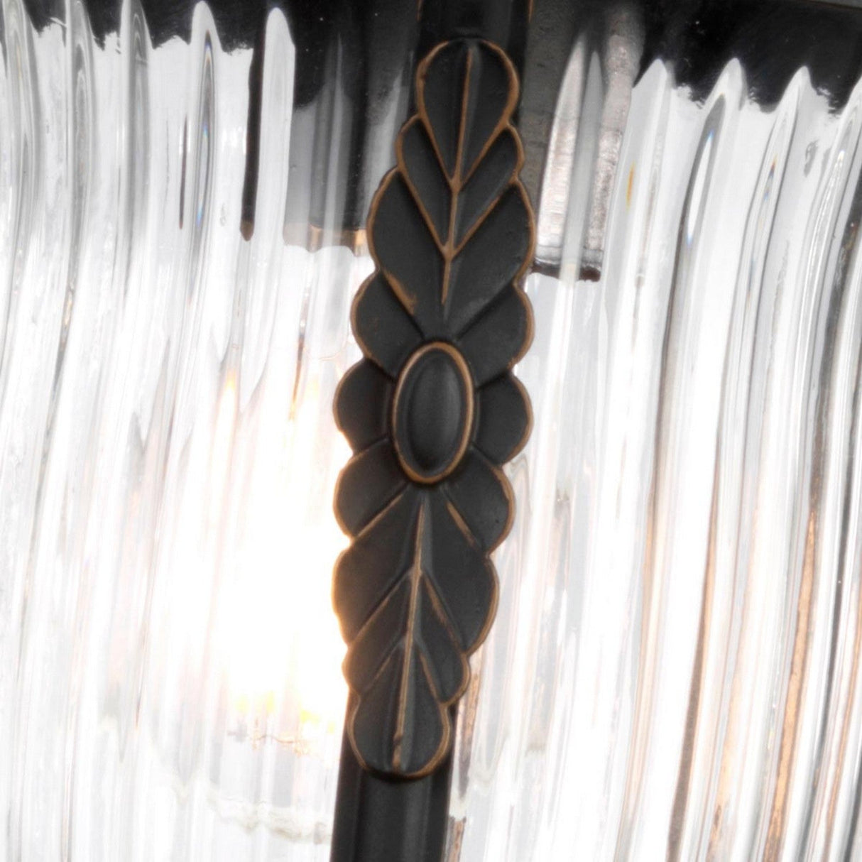 Close-up of a Luverne Outdoor 1 Light Medium Wall Lantern in rubbed bronze, showcasing a central metal embellishment with a leaf pattern and an oval centerpiece. The lantern features a ribbed glass shade that is textured, allowing light to glow softly through it.