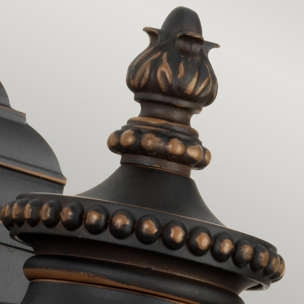 A close-up of the Luverne Outdoor 1 Light Small Wall Lantern in rubbed bronze showcases an ornate black metal finial with bronze accents. The lantern features a flame-shaped top and round beaded details, creating an intricate yet elegant design that enhances its timeless appeal against a plain background.