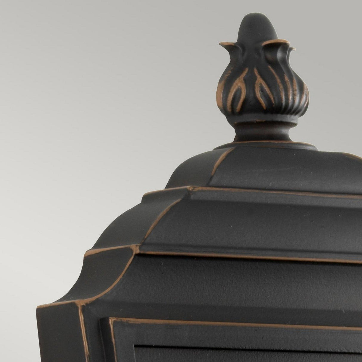 Close-up of a decorative black finial with a flame-like design on the Luverne Outdoor 1 Light Small Wall Lantern - Rubbed Bronze. The smooth finish, accented with subtle rubbed bronze details, is set against a light gray background and evokes elegance with its ribbed glass shade.