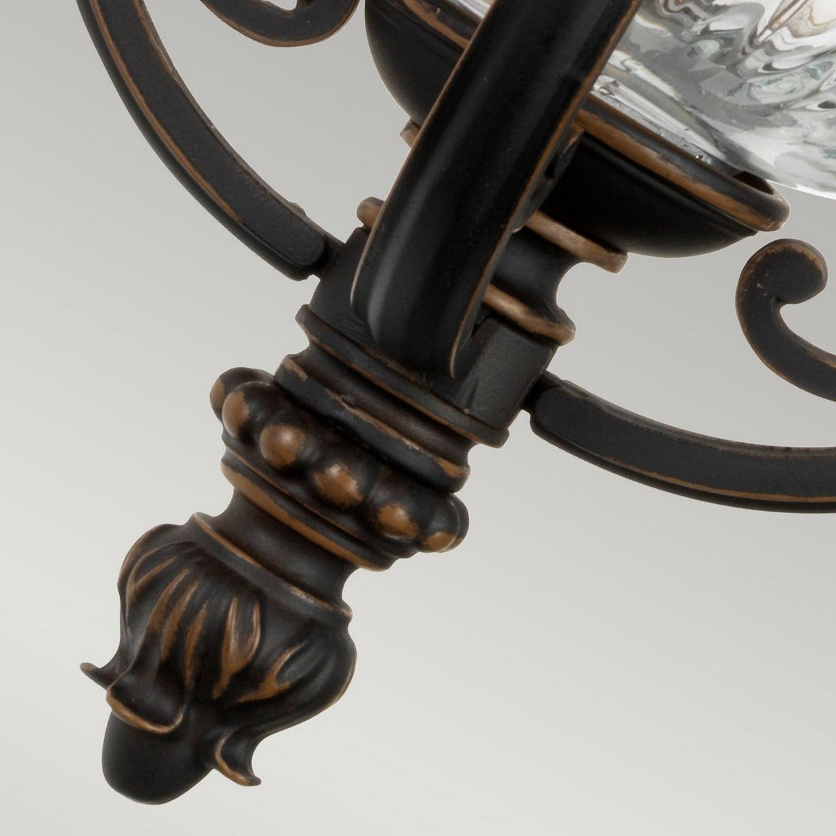 A close-up of the Luverne Outdoor 1 Light Small Wall Lantern reveals its ornate black metal construction with intricate rubbed bronze detailing. The elegant design showcases swirling patterns and a textured central shaft, reminiscent of an antique candle holder, creating a classic appearance against a neutral background.
