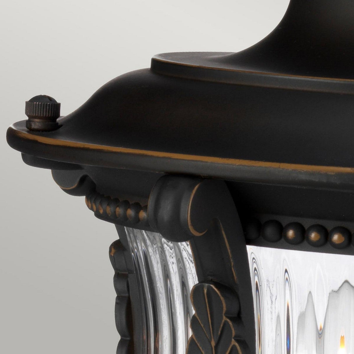 Close-up of the Luverne Outdoor 1 Light Medium Pedestal in rubbed bronze, showcasing its decorative top with ornamental edges and clear, ribbed glass panels. This IP44-rated fixture includes intricate detailing along the frame.