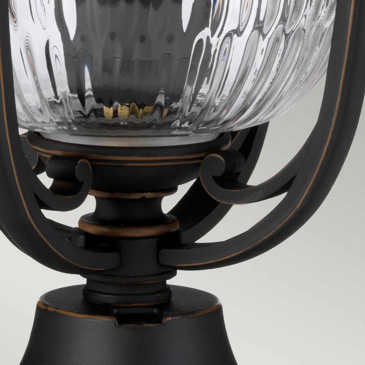 The Luverne Outdoor 1 Light Medium Pedestal in Rubbed Bronze showcases a close-up of its ornate black metal base with a clear, ribbed glass globe. This pedestal light features decorative scroll elements supporting the globe, and its exquisite design is accentuated by the neutral gray background.