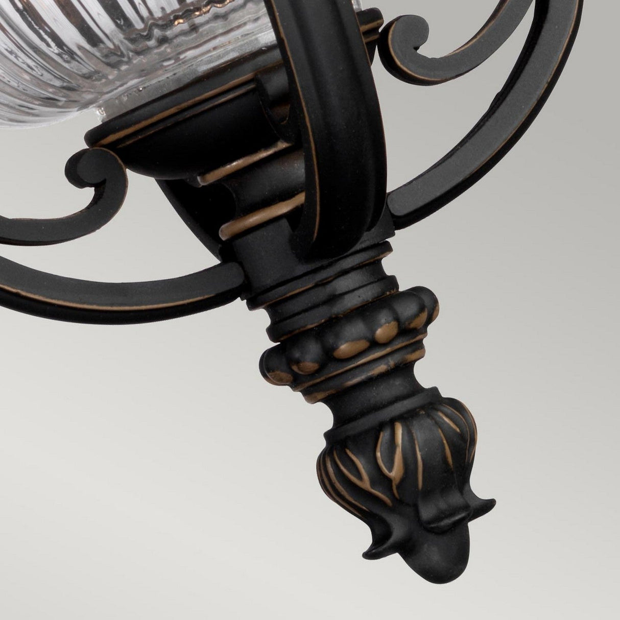 A close-up view of the Luverne Outdoor 1 Light Medium Chain Pendant, showcasing its ornate metal fixture in a rubbed bronze finish. It features a clear ribbed glass shade, complete with intricate curved metalwork beneath it, set against a plain gray background.