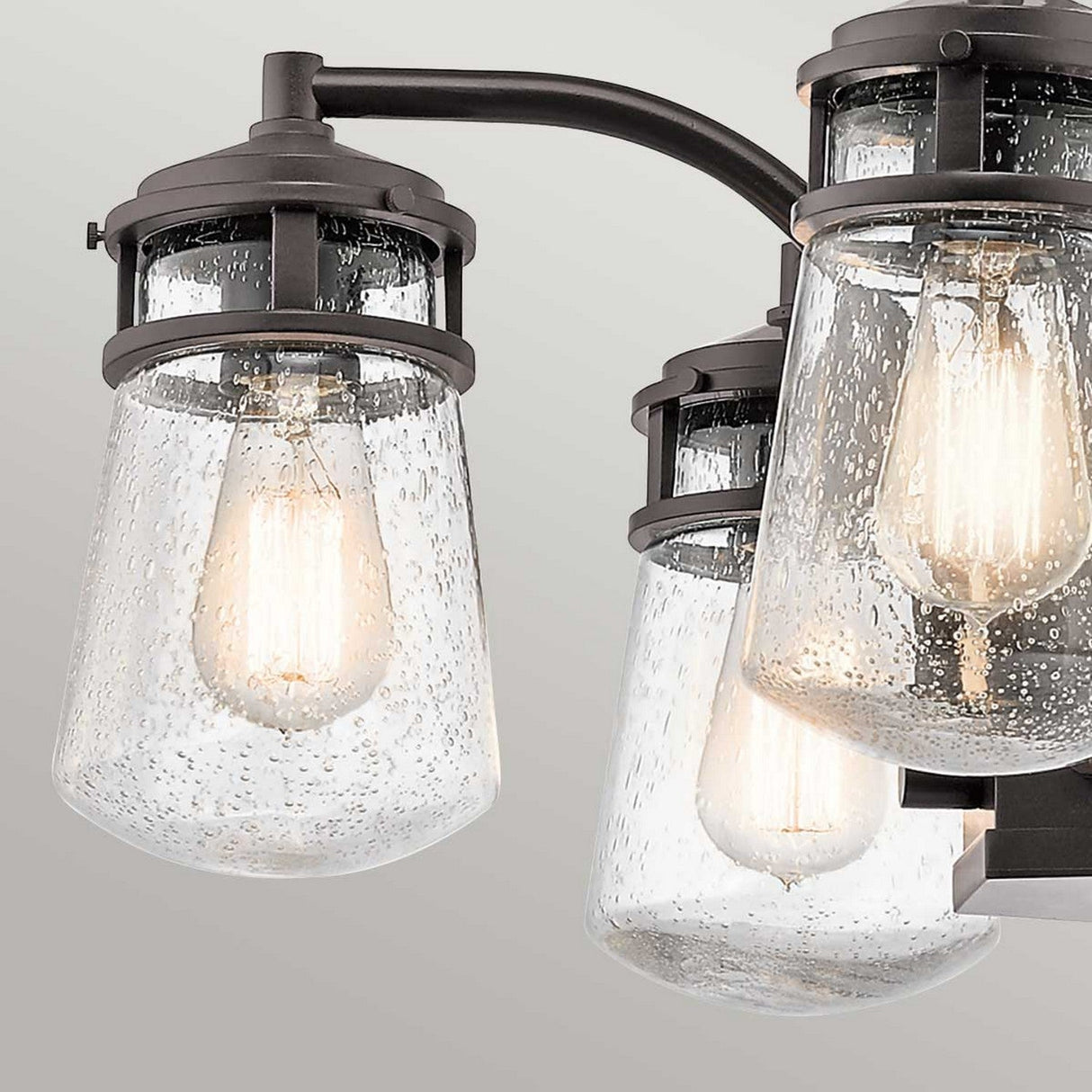 Close-up of the Lyndon Outdoor 5 Light Chandelier showcasing its vintage-style design with a dark bronze metal frame and seedy glass shades encasing three bulbs. The plain, light gray background accentuates its architectural bronze aesthetic, reminiscent of a sophisticated outdoor fixture.