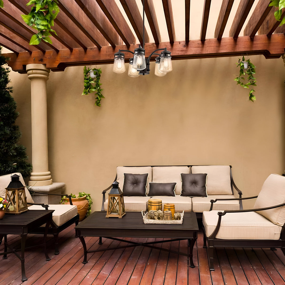 A cozy outdoor patio with a wooden pergola above features beige cushioned seating, a coffee table adorned with lanterns and candles, and decorative greenery hanging. The Lyndon Outdoor 5 Light Outdoor Chandelier in bronze adds a touch of elegance, making the space warm and inviting.