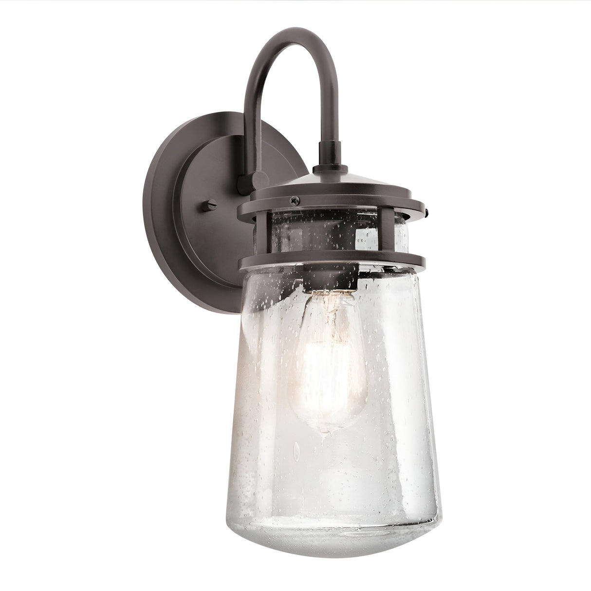 The Lyndon Outdoor 1 Light Medium Wall Lantern in bronze showcases a vintage style with its seedy glass shade. It casts a warm glow through the textured glass and is mounted on a circular backplate, complete with an elegantly curved arm design.