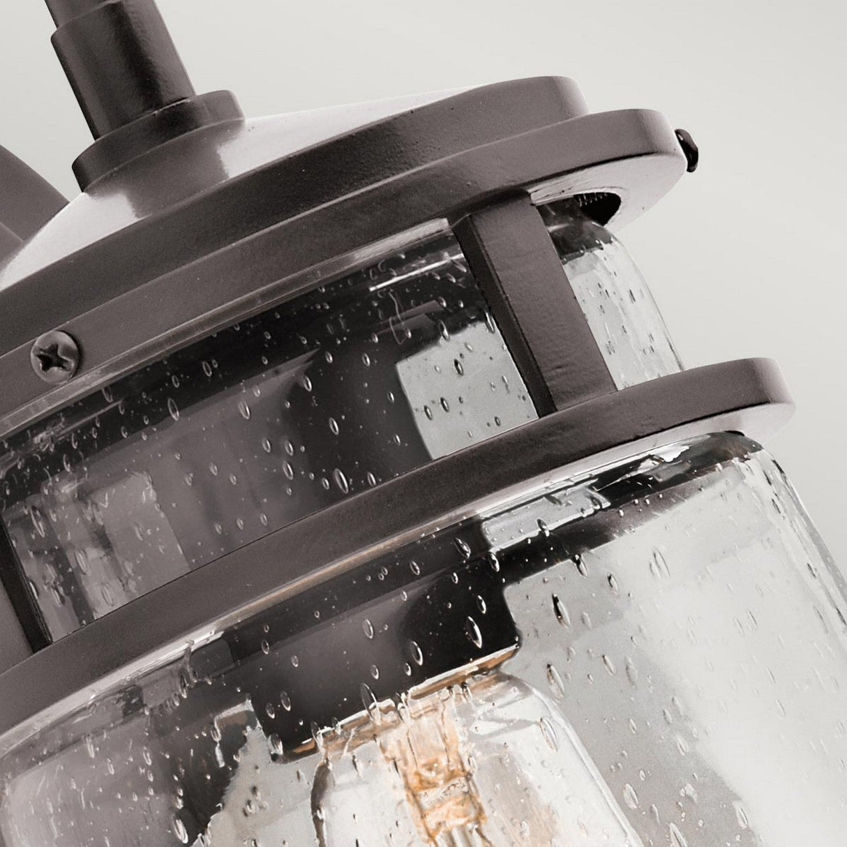 Close-up of the Lyndon Outdoor 1 Light Medium Wall Lantern in bronze, featuring a rustic design with a clear glass cylinder and black metal frame. The glass surface has water droplets, hinting at recent rain. The fixture is illuminated, casting a warm light through its seedy glass shade.