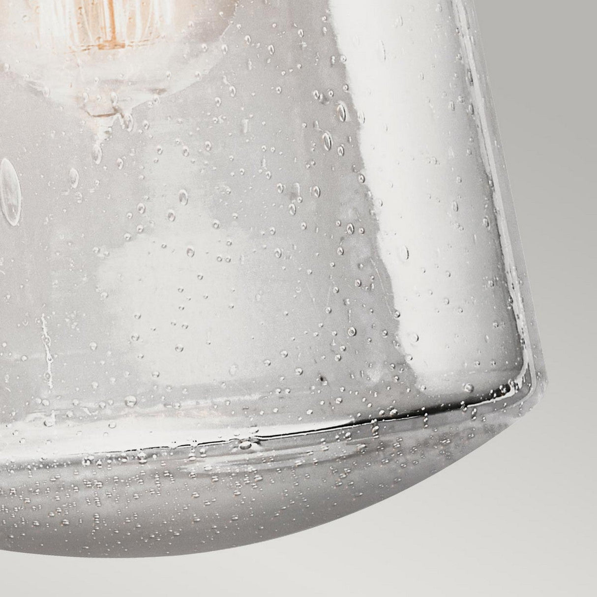 Close-up of a clear seedy glass shade on the Lyndon Outdoor 1 Light Medium Wall Lantern - Bronze, showcasing its textured surface with delicate bubbles. Soft light filters through, with a neutral gray background enhancing the elegance of the design, echoing the charm of an outdoor wall lantern.