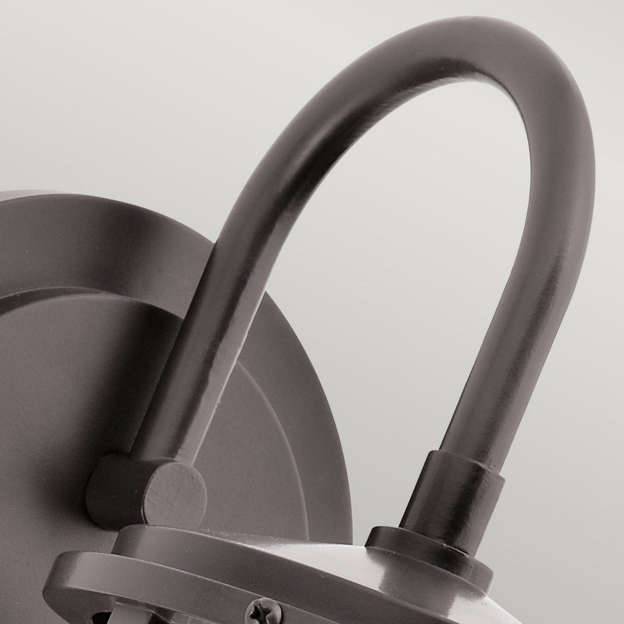 A close-up of the Lyndon Outdoor 1 Light Medium Wall Lantern reveals its modern design with a sleek, curved arm in a dark bronze finish. The lantern's minimalistic style showcases simple lines and a seedy glass shade for added texture, all secured by a screw at the base.