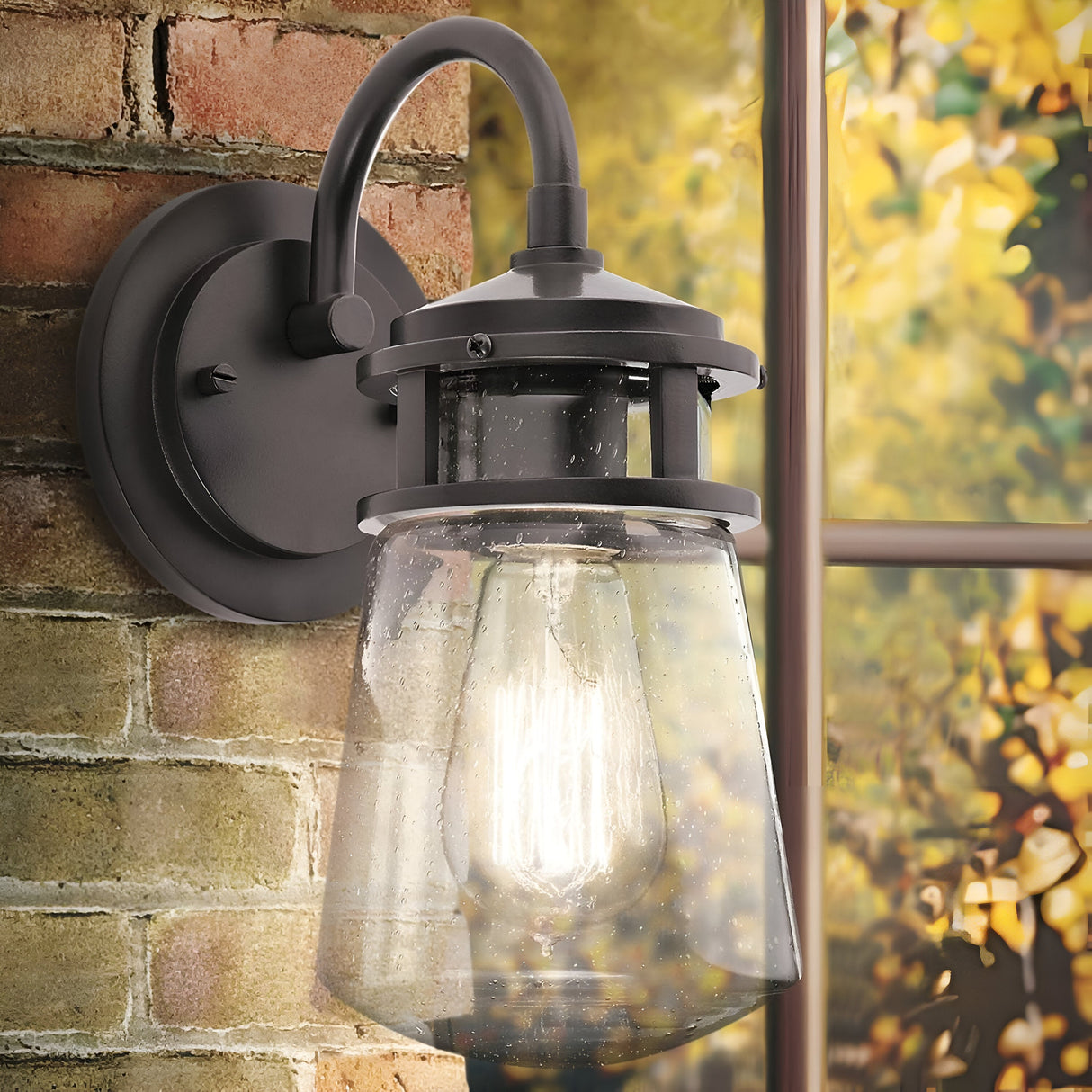 The Lyndon Outdoor 1 Light Medium Wall Lantern in bronze is installed on a brick wall near a window, showcasing a vintage-style aesthetic. It features a robust metal frame and seedy glass shade, casting a warm glow that beautifully illuminates the blurred autumn foliage visible through the window.