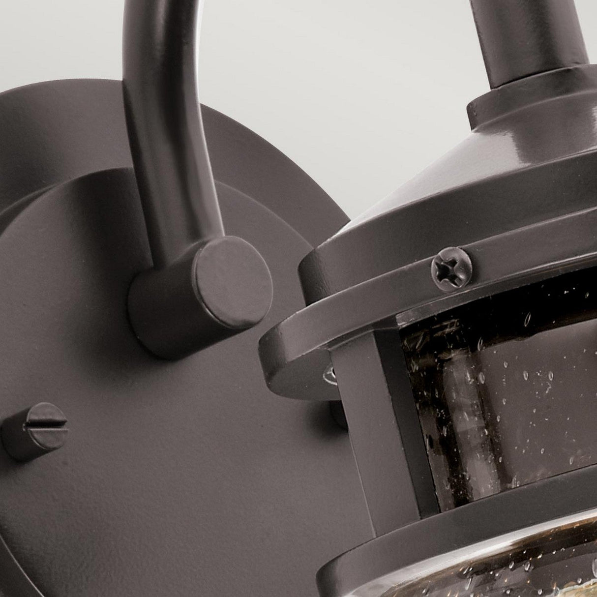 Close-up of the Lyndon Outdoor 1 Light Small Wall Lantern in bronze, showcasing its clear seedy glass panels. The fixture is mounted on a wall with visible screws and features a curved arm. The image emphasizes the top half of the lantern, highlighting its modern design.