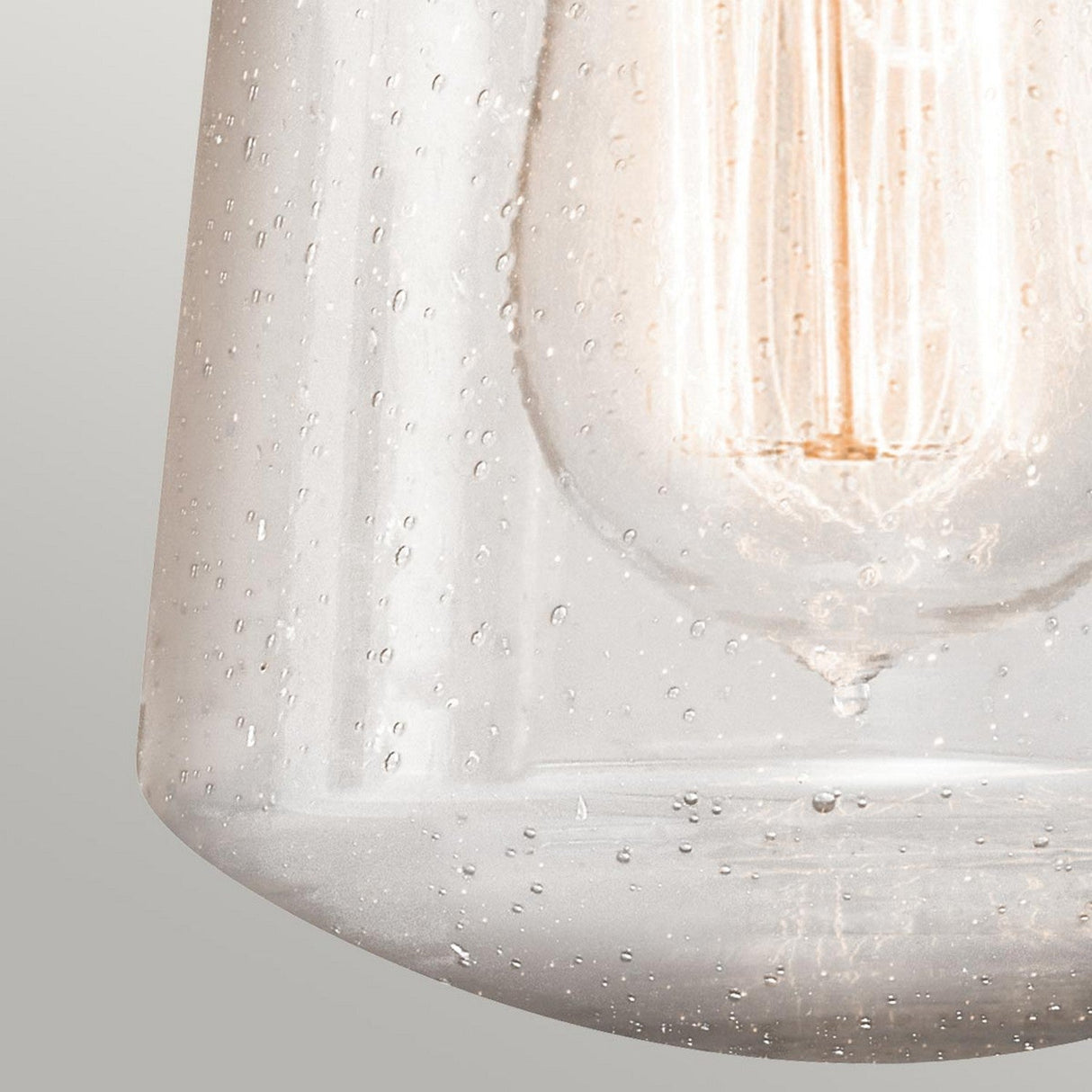 A close-up view of the Lyndon Outdoor 1 Light Small Wall Lantern in bronze showcases its clear seedy glass design adorned with small air bubbles. Inside, a filament bulb casts a warm glow, creating an ambiance on a plain, neutral background that enhances its outdoor lantern aesthetic.