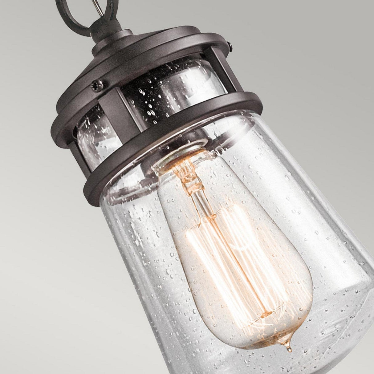 Close-up of the Lyndon Outdoor 1 Light Small Chain Pendant - Bronze, which showcases a vintage-style design with a seedy glass shade featuring a water droplet texture. It houses an Edison-style bulb that emits a warm glow and is mounted on an architectural bronze frame with a dark finish, set against a light background.