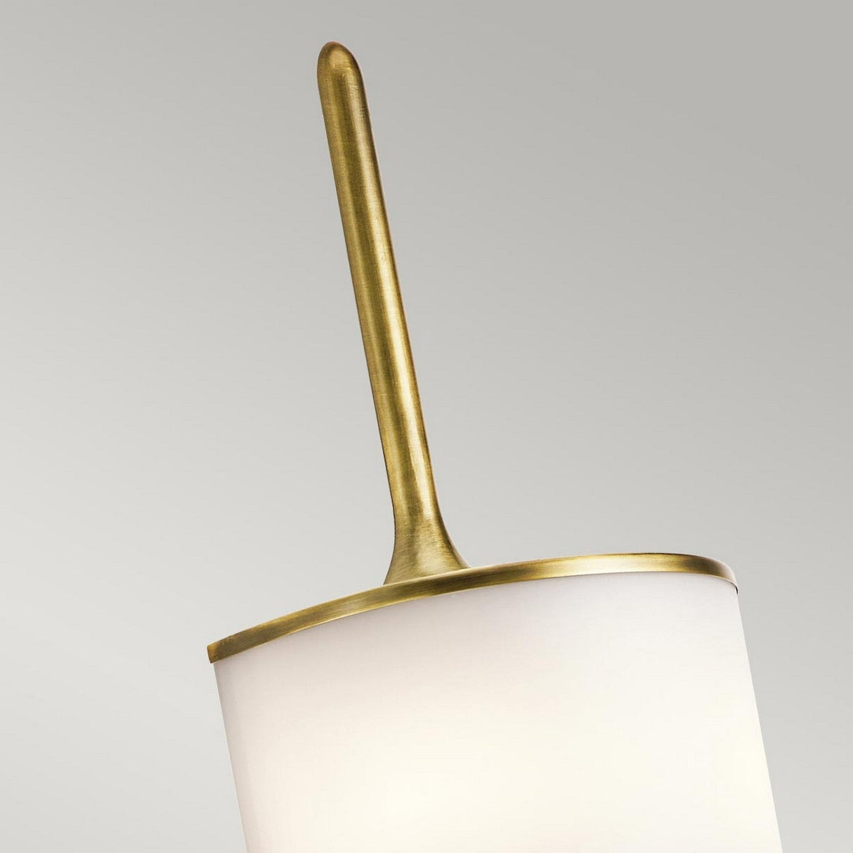 A close-up of the Mona 2 Light Large Wall Light - Natural Brass showcases its elegant white cylindrical shade and natural brass accent at the top. The sleek rod extends upwards, emitting light from energy-efficient LEDs, all set against a neutral gray background.