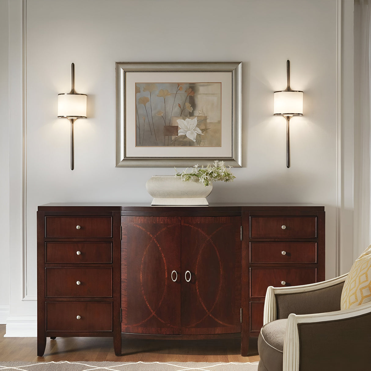 The room boasts a wooden sideboard with six drawers and two central cabinets, topped with a vase of flowers. Above it hangs a framed floral painting, complemented by elegant Mona 2 Light Small Wall Lights in a sleek silver finish. On the right, an armchair is partially visible.