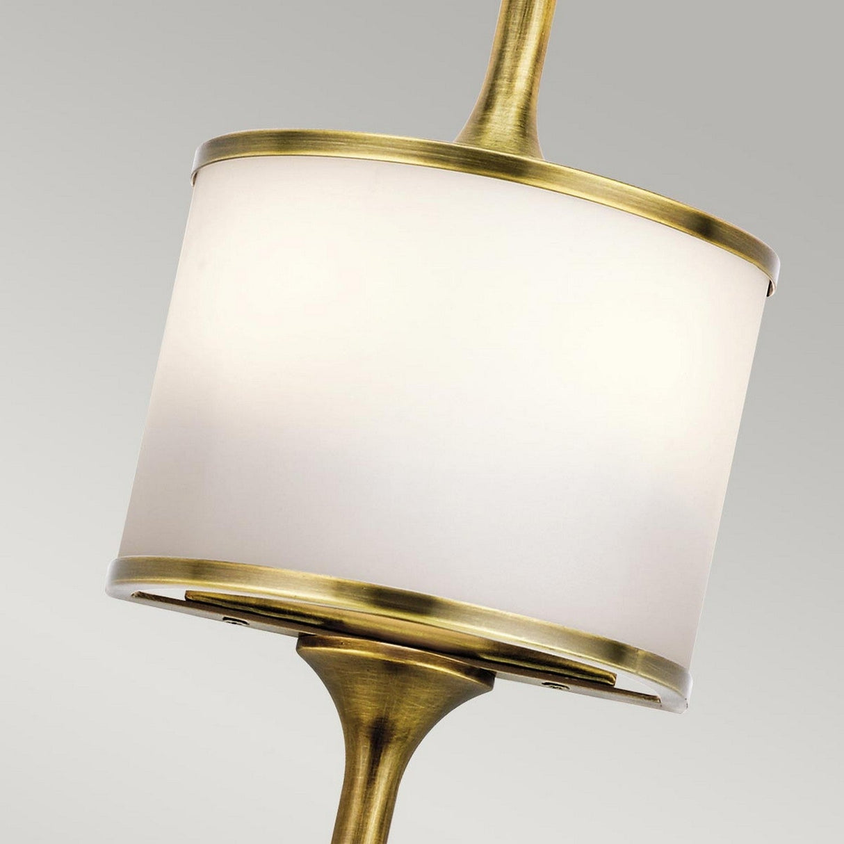 A close-up of the Mona 2 Light Small Wall Light in natural brass, highlighting its sleek cylindrical white lampshade. The elegant, mid-century style design is angled beautifully against a neutral gray background.