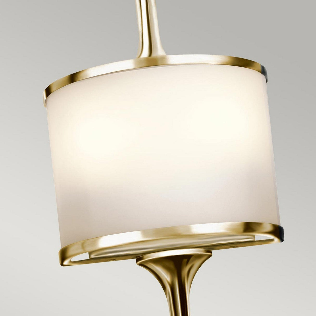 The Mona 2 Light Small Wall Light, featuring a cylindrical white shade and sleek polished brass accents, is displayed against a plain gray background.