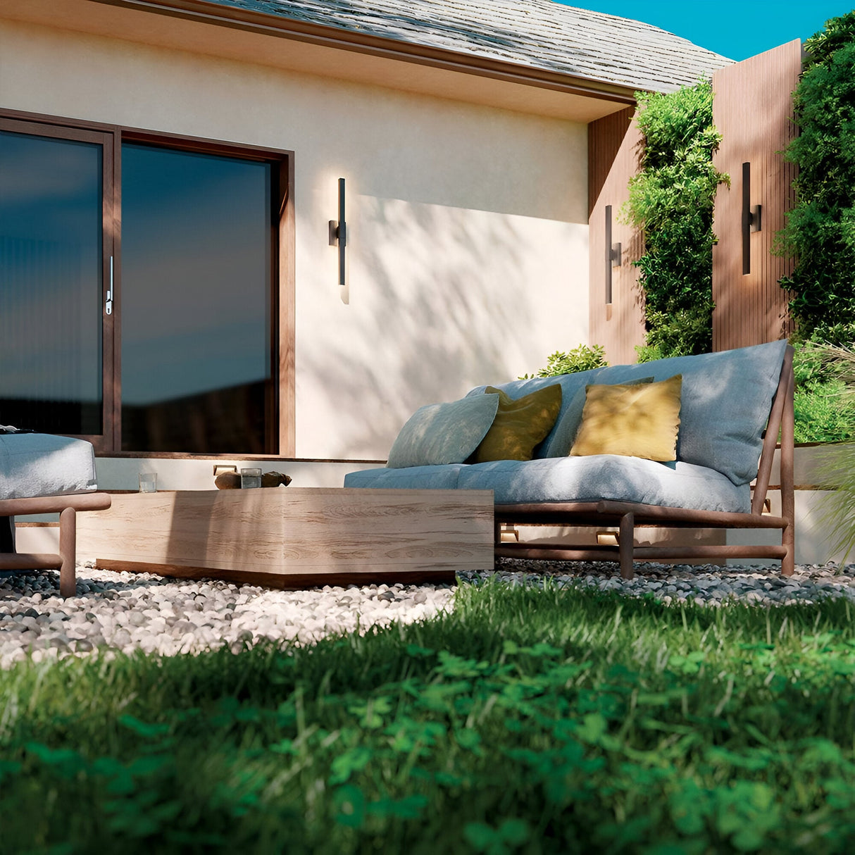 Modern outdoor patio with cushioned wooden lounge chairs, yellow and gray pillows, a low wooden table, gravel ground, green plants, and a beige house featuring large glass doors with the Nocar LED Outdoor Large Wall Light 39W 3000K in black.