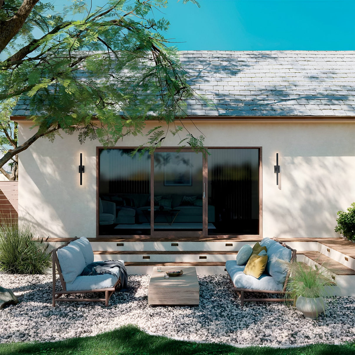 A modern patio with two cushioned lounge chairs, a low wooden table, decorative pillows, and potted plants on gravel is set in front of a house featuring Nocar LED Outdoor Large Wall Light 39W 3000K - Black and large sliding glass doors.