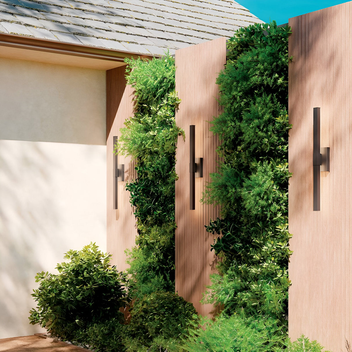 Vertical wooden panels showcase lush green plants in a modern garden. The Nocar LED Outdoor Large Wall Light 39W 3000K in black delivers stylish exterior lighting. A building’s roof and clear blue sky are also visible.