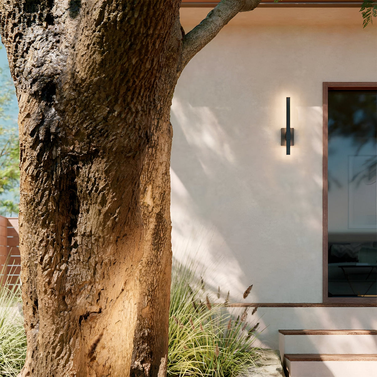 A large tree trunk partially shades a modern house exterior, which features a Nocar LED Outdoor Medium Wall Light 20W 3000K - Black as a minimalist sconce, plus a glass door and ornamental grasses by the entrance steps.