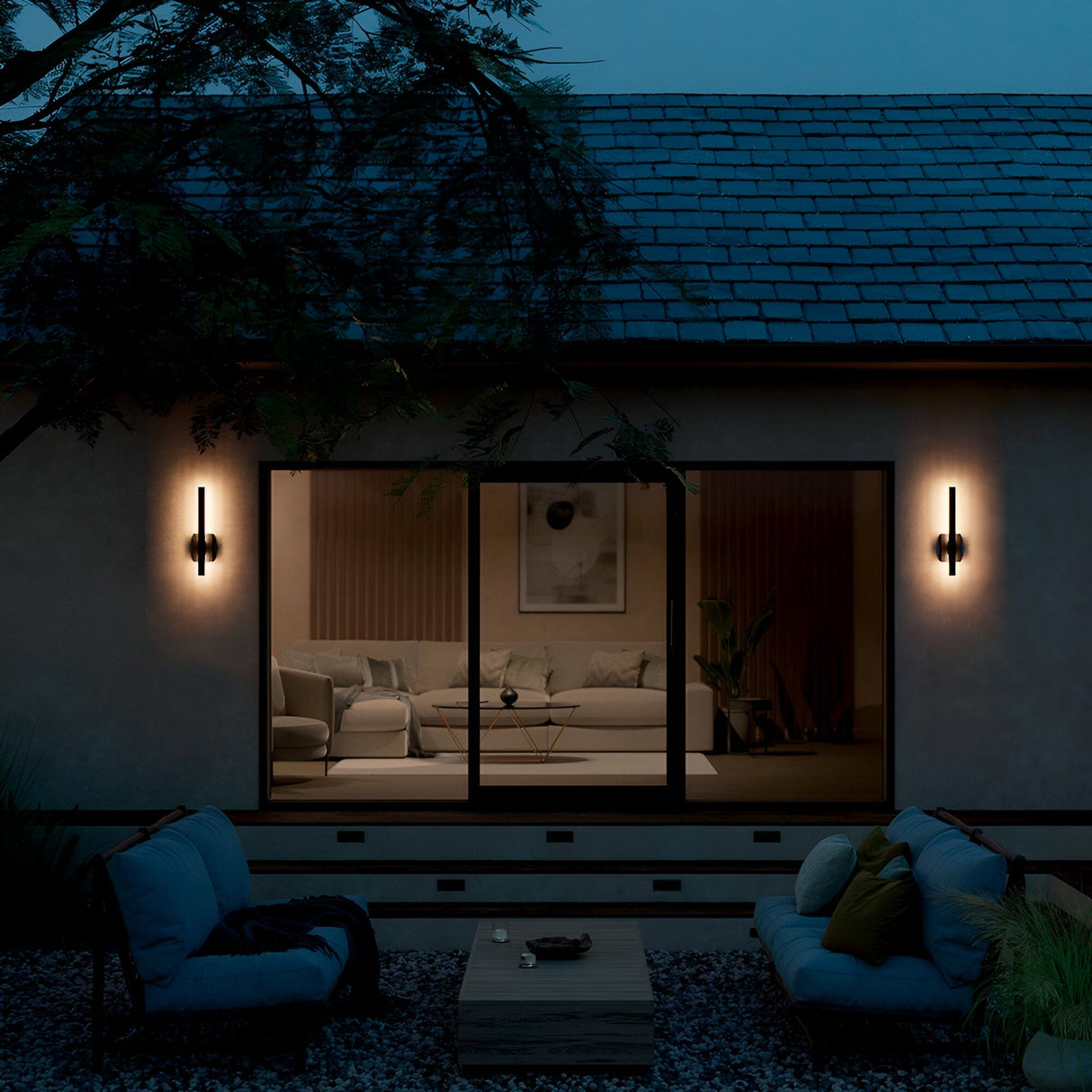 A modern house at dusk features Nocar LED Outdoor Medium Wall Light 20W 3000K - Black flanking large glass doors; inside, a bright living room with sofa and plants awaits. The patio showcases modern lighting, cushioned chairs, a low table, and nearby trees.