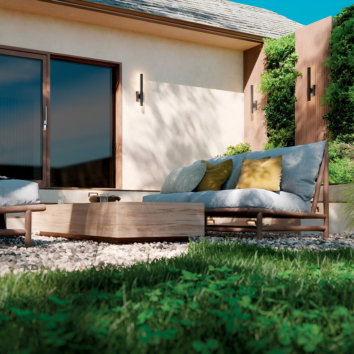 A modern patio with cushioned wooden lounge chairs, green and yellow pillows, a low wooden table, gravel ground, lush grass, and a beige building featuring large glass doors illuminated by Nocar LED Outdoor Medium Wall Light 20W 3000K - Black.