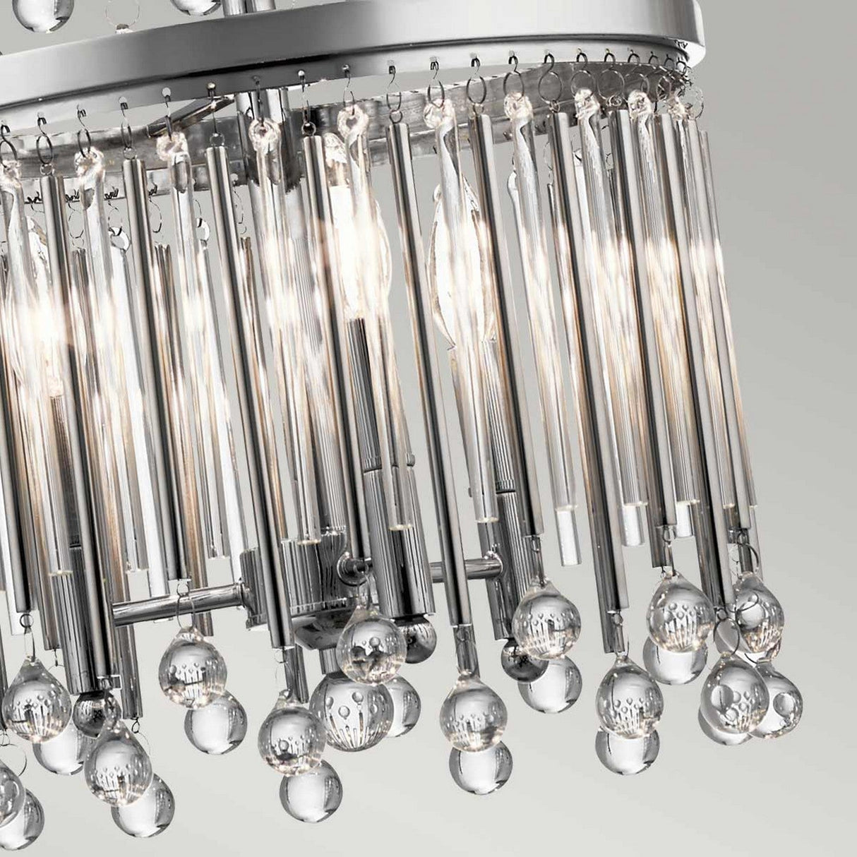 Close-up of the Piper 18 Light Chandelier in polished chrome, showcasing its modern design with glass bead accents at the bottom. The chrome rods encircle the light bulbs, producing a luxurious and sparkling effect against a soft gray background.