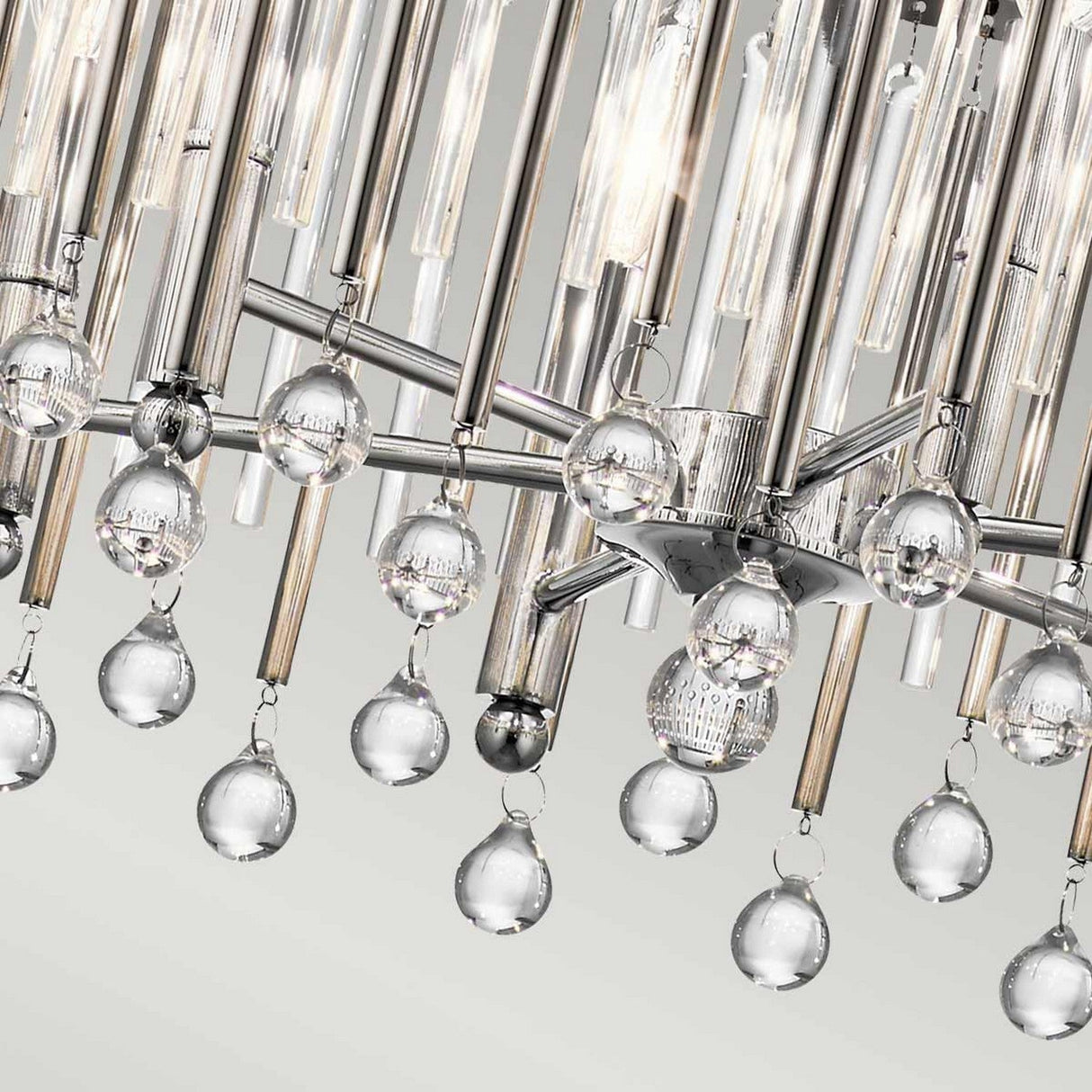 A detailed look at the Piper 6 Light Chandelier - Polished Chrome showcases its sleek chrome frame embellished with hanging glass rods and round glass droplets, while the glass bead accents enhance its elegance by reflecting light beautifully against a pristine, light grey backdrop.