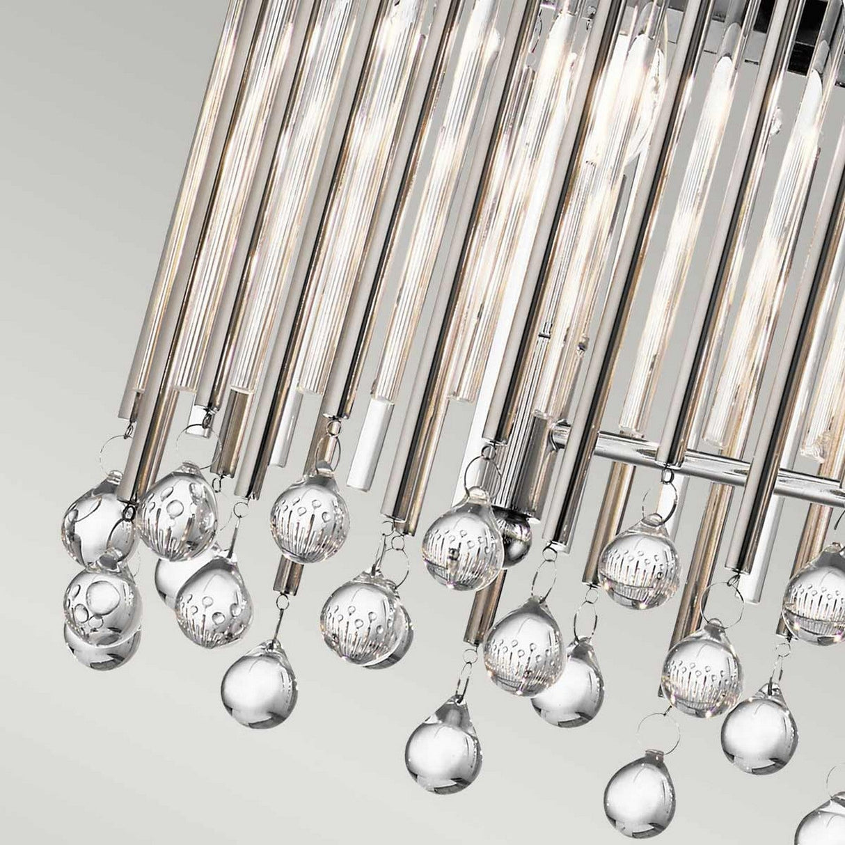 A close-up of the Piper 6 Light Oval Chandelier reveals its elegant design, featuring glass bead accents and an array of hanging clear globes with elongated cylindrical lights. The polished chrome elements enhance its sophistication, adding a contemporary touch against a neutral background.