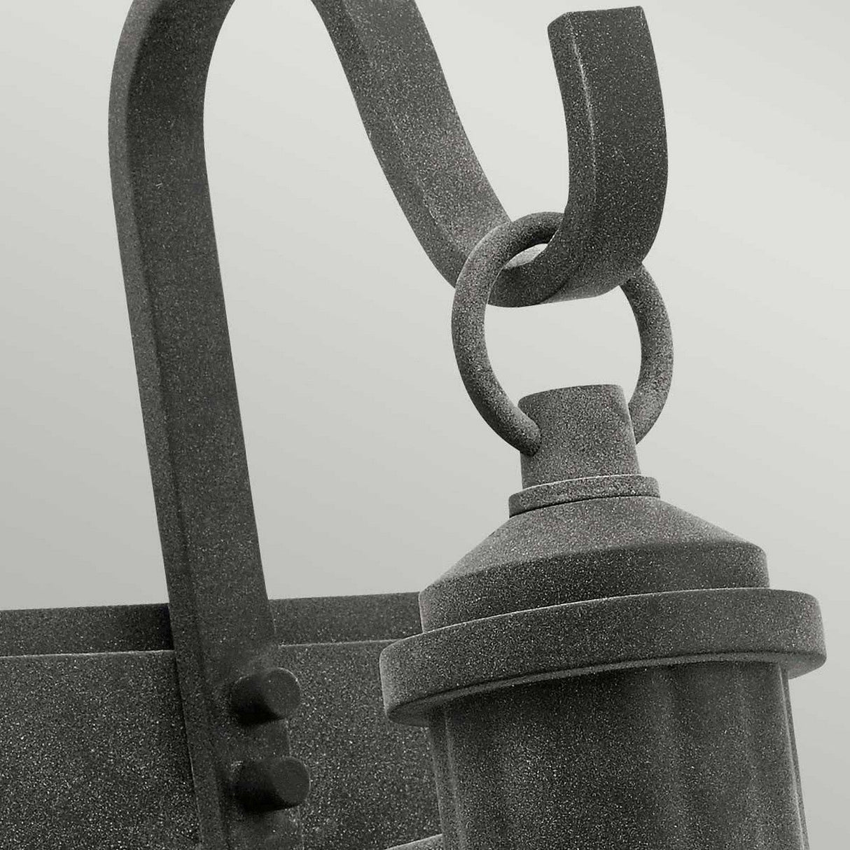 Close-up image of the Riverwood Outdoor 1 Light Medium Wall Lantern, highlighting its rustic elegance with a weathered zinc finish. The design showcases a matte-finished curved metal hanger and cylindrical light fixture against a light gray background.
